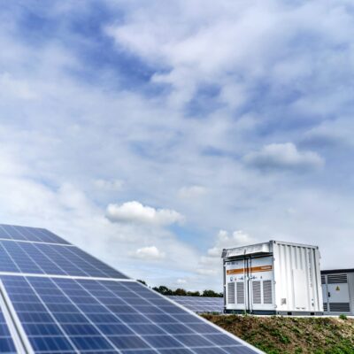 Rows of blue solar panels in a solar farm under a partly cloudy sky, with industrial equipment in the background.