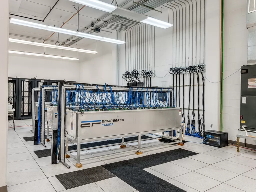 Industrial lab with a stainless steel modular unit labeled 'Engineered Fluids' and blue tubing, network racks, and cables in a clean white room