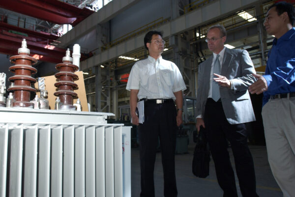 Three engineers in a factory inspect large brown electrical insulators mounted on a cabinet in an industrial setting.