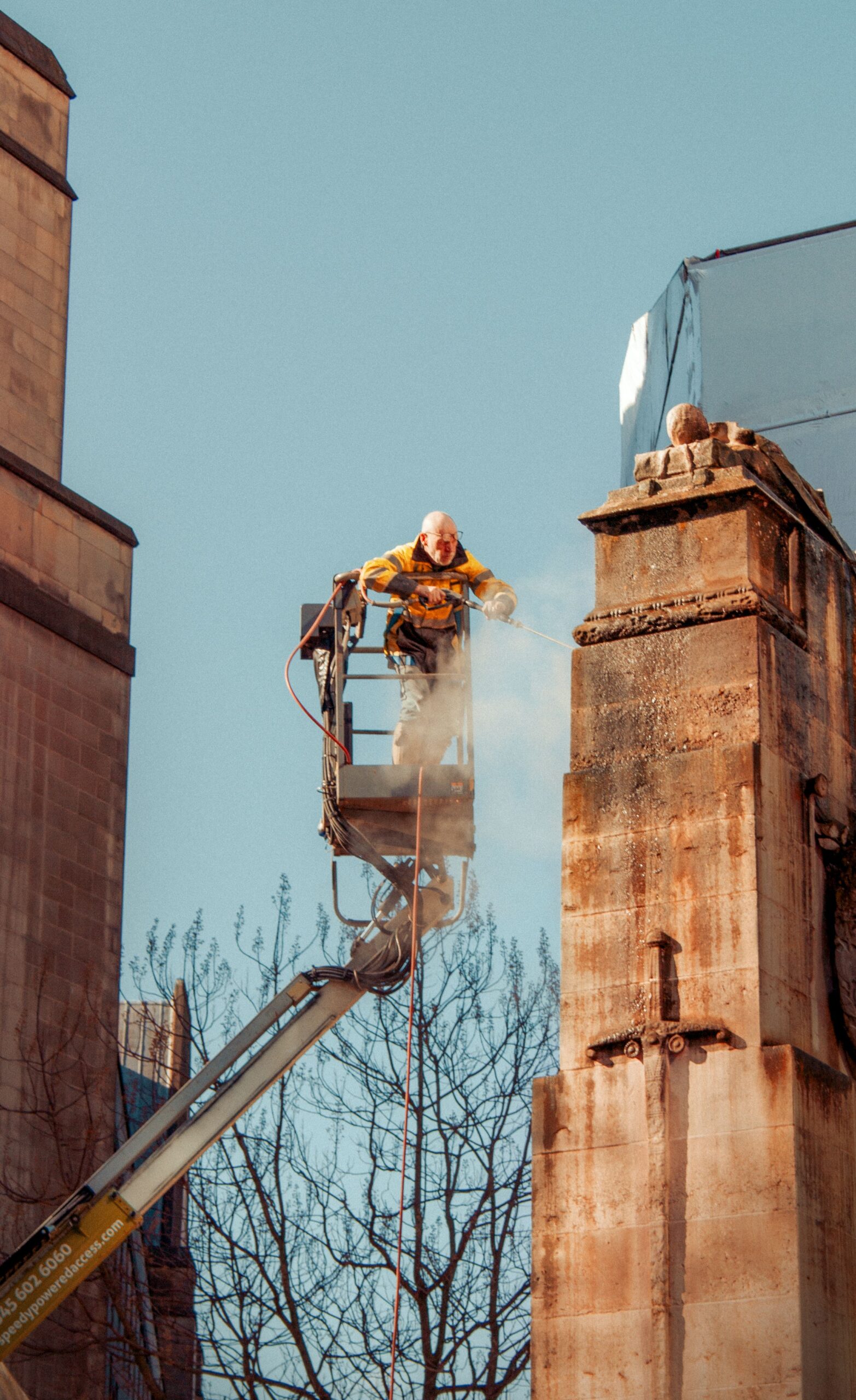Worker in a yellow safety jacket on a raised platform cleaning a tall stone column between buildings with a clear blue sky behind him.