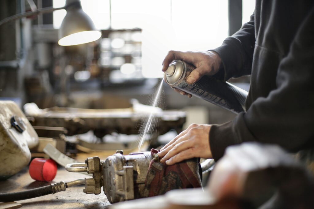Person using a spray can to apply liquid onto a workpiece at a cluttered workshop bench