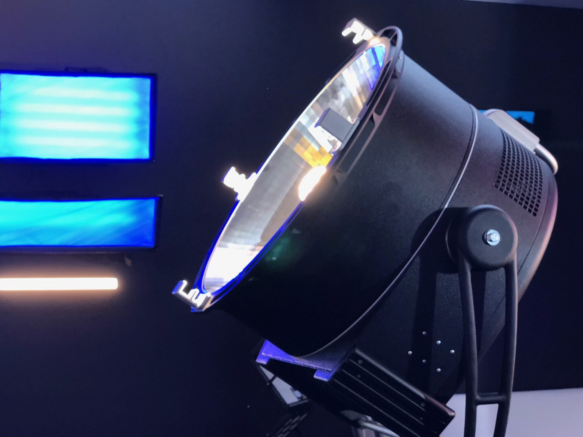 Close-up of a large black studio microphone on a stand in a dark room with blue-lit screens in the background.